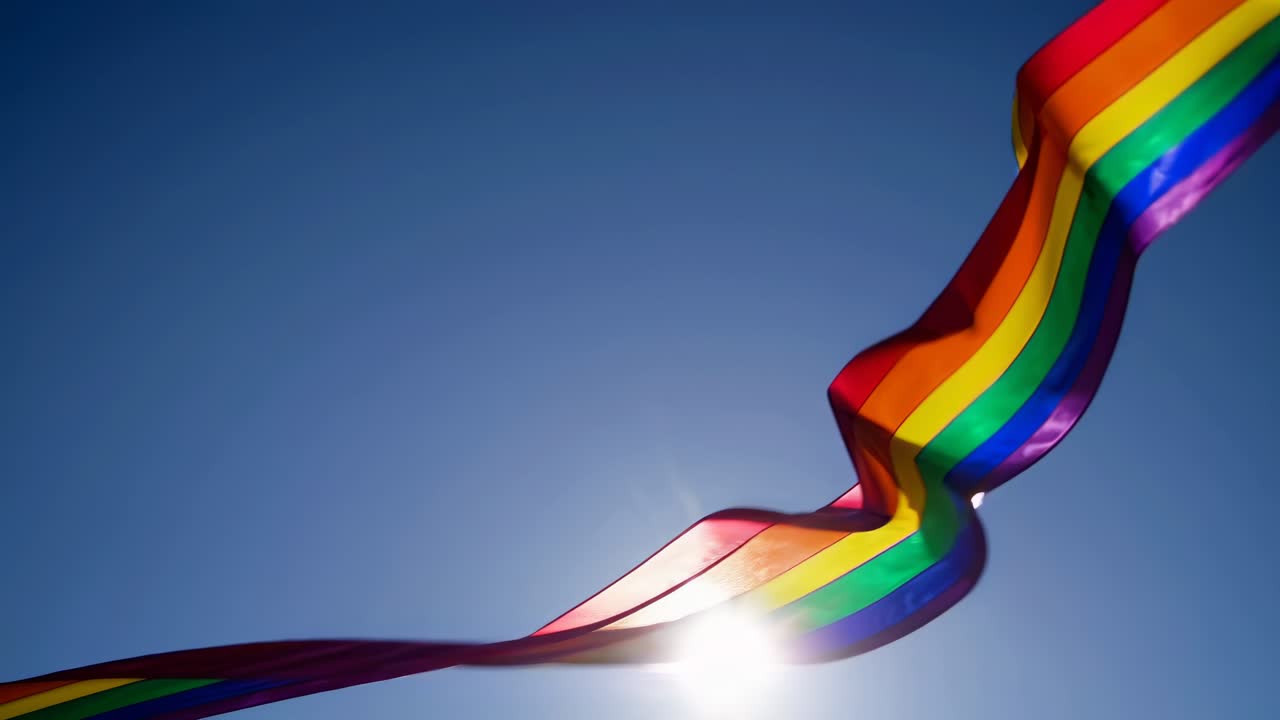 Low-angle video shot of colorful ribbons and a rainbow flag waving against a clear blue sky