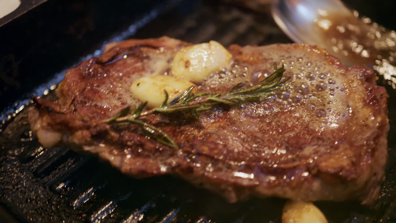Ein Steak brät in einer Grillpfanne und wird dabei mit geschmolzener Butter und Kräutern übergossen.