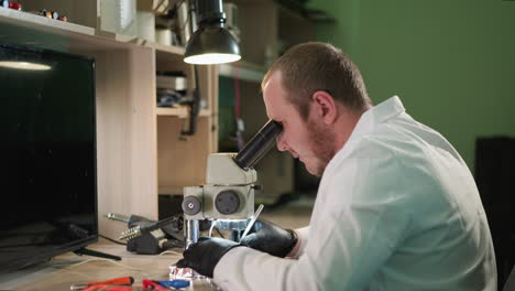 un técnico enfocado en una bata blanca de laboratorio recoge algo en la mesa y está trabajando en el circuito bajo un microscopio en un laboratorio bien equipado, con varios equipos en el escritorio