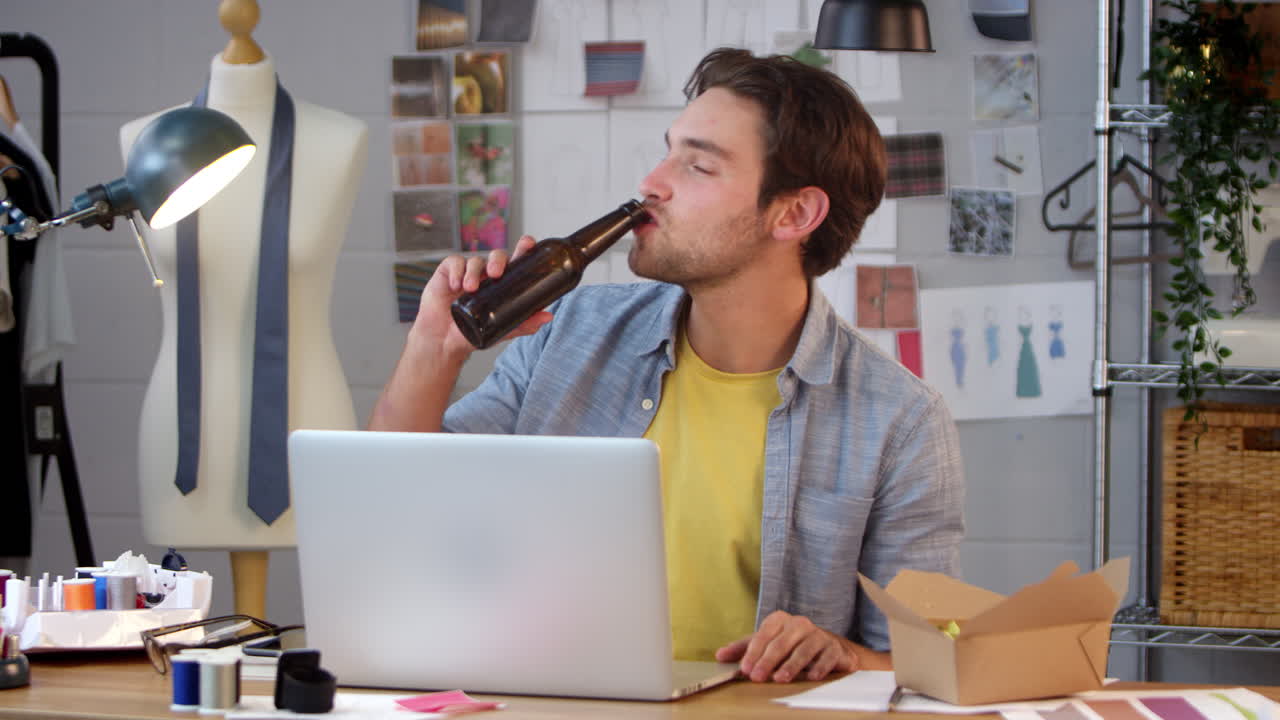 propietario masculino de un negocio de moda con una botella de cerveza escuchando música en una computadora portátil en el estudio