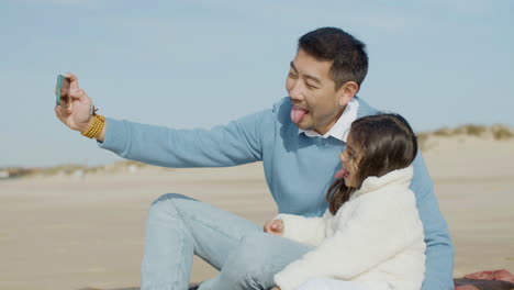 Japanischer-Vater-Und-Tochter-Machen-Selfie-Mit-Dem-Handy-Und-Zeigen-Zungen,-Während-Sie-Gemeinsam-Zeit-Am-Strand-Verbringen