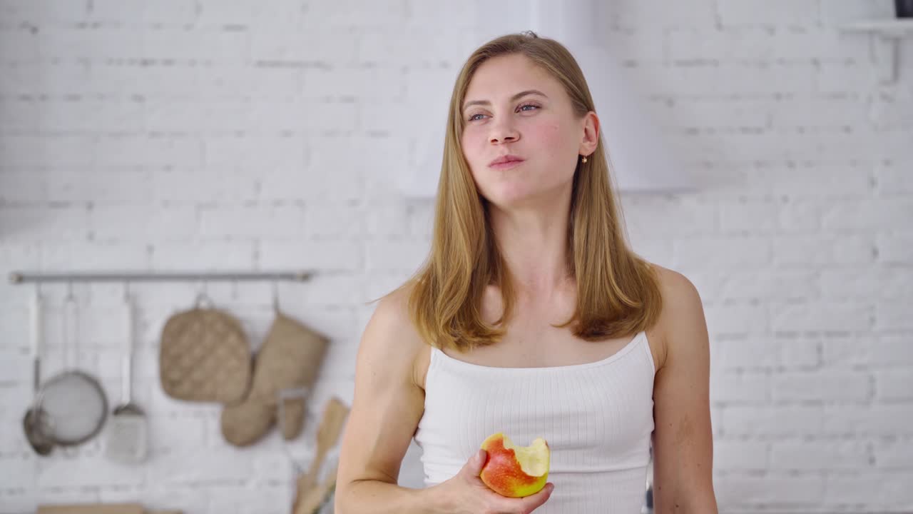 Fit smiling young woman eating healthy fruit. Beautiful girl enjoys eating fresh sweet apple on the modern kitchen background. Concept of losing weight.