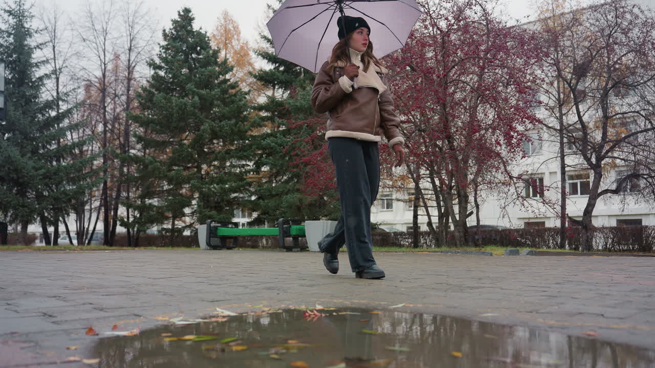 Une femme se promène paisiblement dans un parc pavé par une froide journée nuageuse, tenant un parapluie, vêtue d’un bonnet en tricot noir, d’une veste en peau de mouton marron et d’un pantalon noir, entourée d’arbres à feuilles persistantes et de légères chutes de neige