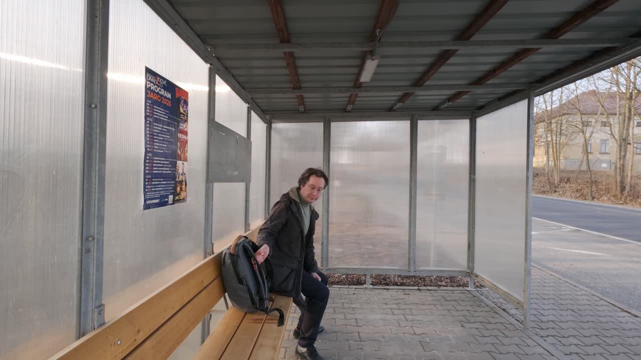 Uomo seduto su una panchina alla fermata dell'autobus che controlla l'ora prima di partire. Scena urbana che cattura il momento dell'attesa e della partenza.