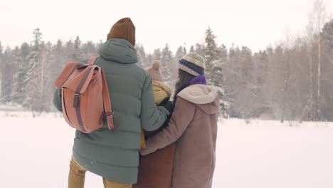 rückansicht von vater, mutter und tochter, die sich umarmen, während sie die landschaft in einem verschneiten wald betrachten