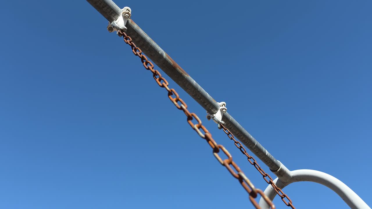 Catene dell'altalena usurate e arrugginite cigolano mentre oscillano avanti e indietro. Il metallo ossidato color marrone-arancio crea un contrasto netto e materico contro il blu intenso e senza nuvole di un ampio cielo del Midwest.