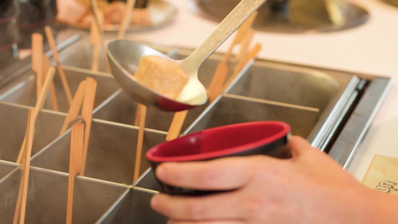 Close-up of a person ladling steaming soup and fish cake skewers into a bowl.