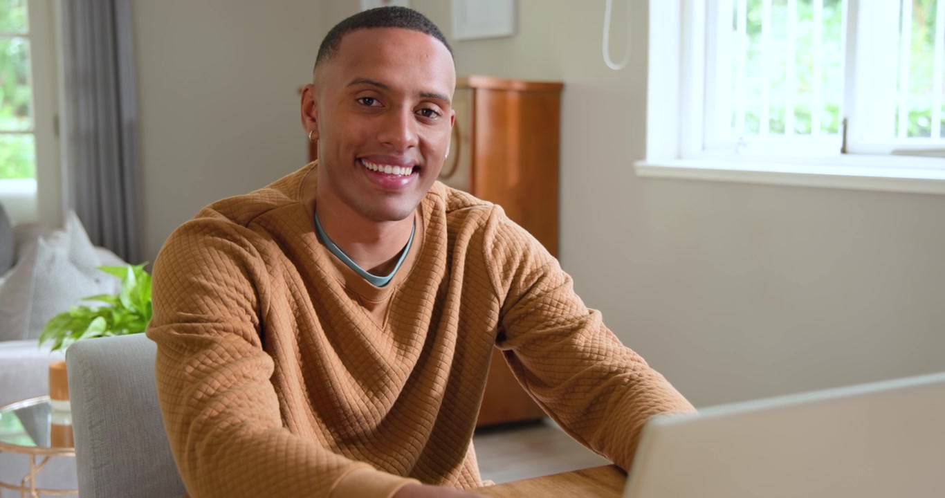 Uomo afroamericano sulla ventina inoltrata che inizia a lavorare da casa, digita sul laptop con una tazza di caffè e sorride.
