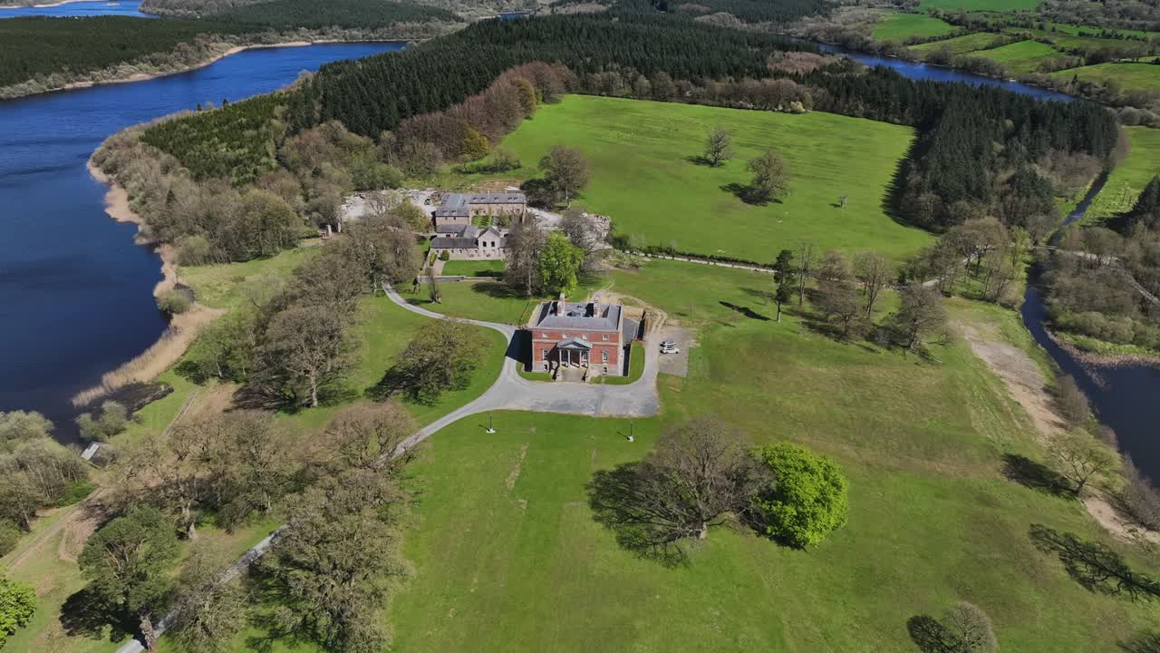 Bellamont House, County Cavan, Ireland, April 2023. Drone orbits clockwise historical architecture surrounded by forest, green fields and lake on sunny day.