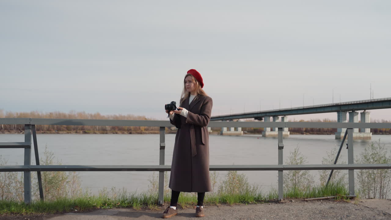 La fotografa immortala il fiume. Donna regola la fotocamera sul ponte. Scatto spontaneo di donna vicino al fiume con fotocamera. Scena autunnale di donna che scatta fotografie spontanee del ponte sul fiume indossando un berretto rosso.