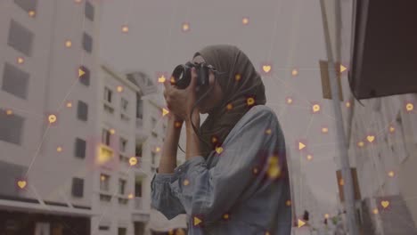 Animation-of-network-of-connected-icons-and-woman-wearing-hijab-taking-photos-in-street
