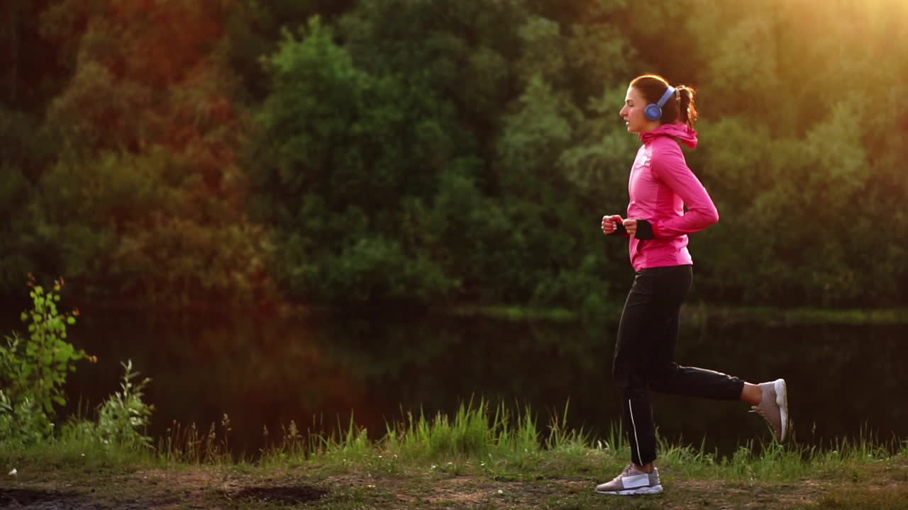 La ragazza corre al tramonto nel parco lungo lo stagno e ascolta musica in cuffia.