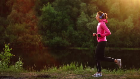 La-Niña-Corre-Al-Atardecer-En-El-Parque-A-Lo-Largo-Del-Estanque-Y-Escucha-Música-Con-Auriculares