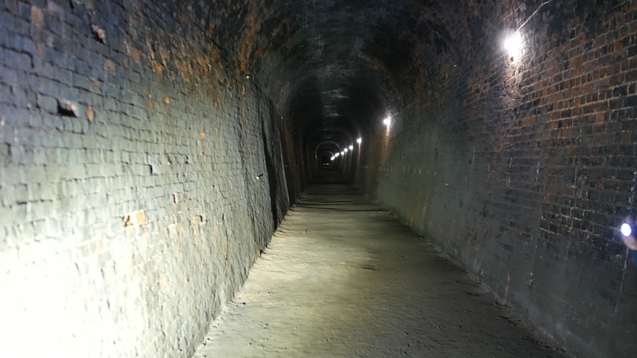 A long, brick-lined tunnel underground with a slightly curved ceiling is illuminated lights along the walls. The dark end of the tunnel, highlighting its depth and structure