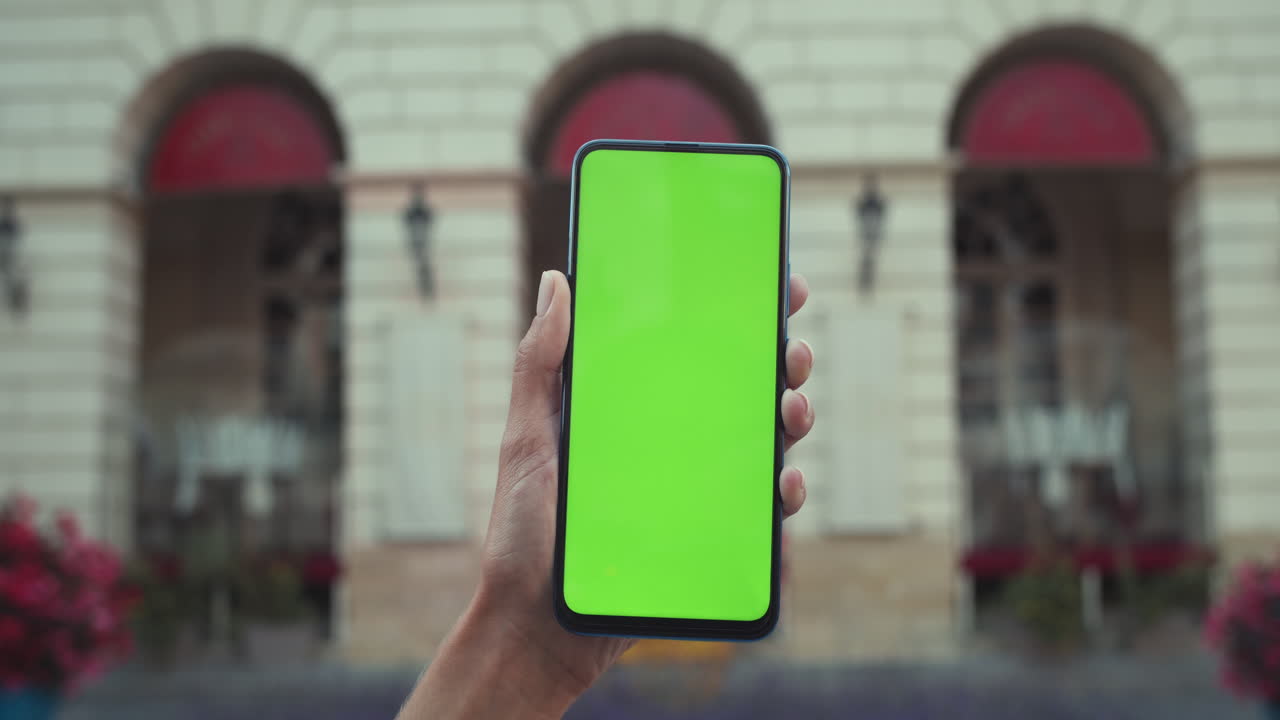 Hand holding a smartphone with a green screen in front of a building