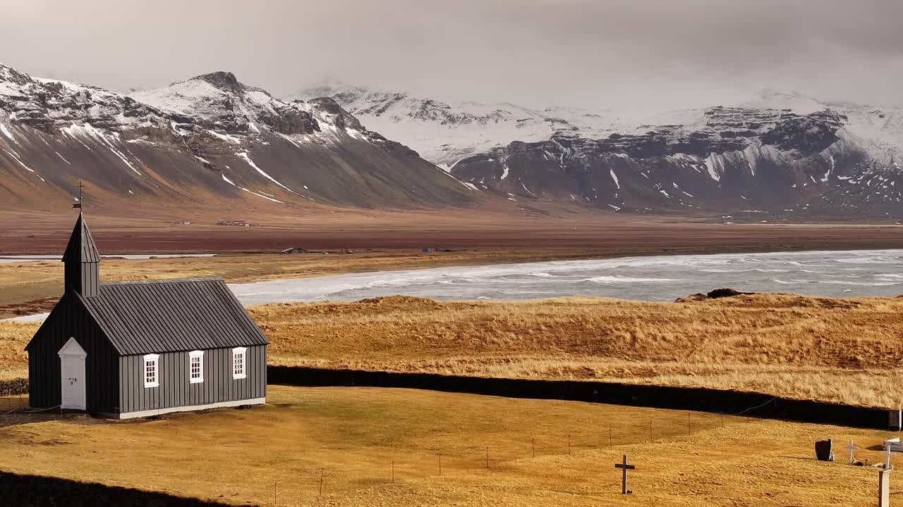 The iconic black Búðakirkja church stands alone in a golden field with snow-capped mountains in the background on the Snæfellsnes Peninsula.