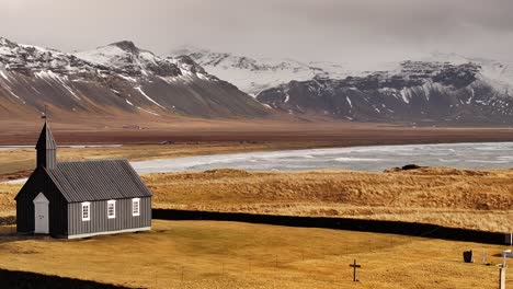 the iconic black búðakirkja church stands alone in a golden field with snow-capped mountains in the background on the snæfellsnes peninsula.
