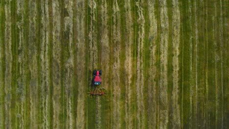 top down aerial drone footage of a small red tractor collecting freshly cut silage hay on a grassy green farm field with cloud and sun patches going over it, rotating paddles collect hay in the back
