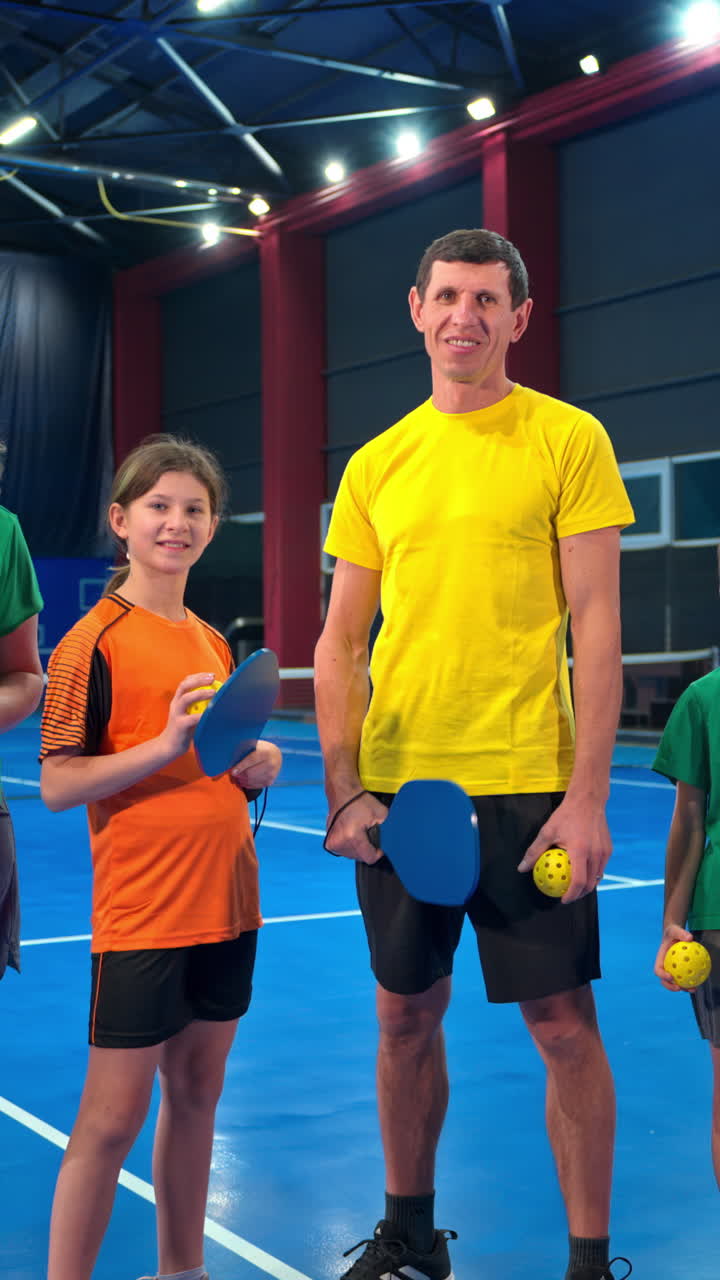 Two adults and two children posing after playing pickleball on a blue, inside court. Vertical