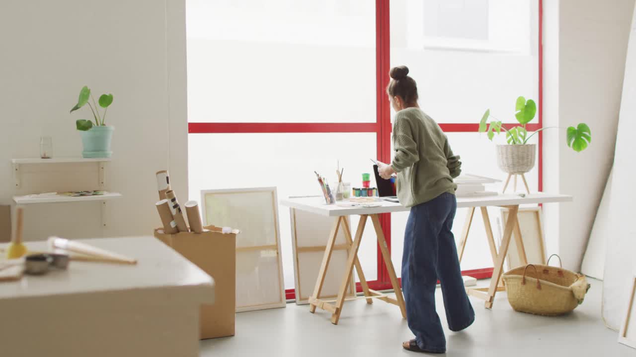 video de una feliz diseñadora biracial usando una computadora portátil y una tableta en el estudio