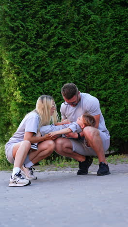 caucasian parents sitting squatted with their child. mom tickles her son and dad kisses him lovingly. vertical video