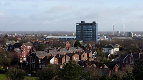 Aerial-view-over-park-trees-to-industrial-rural-townscape-with-blue-skyscraper,-Merseyside,-England