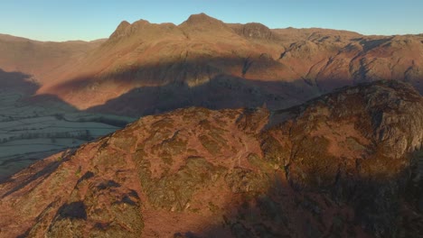 flying towards and over craggy mountain shoulder bathed in early morning winter light revealing deep shadowed valley floor. langdales, english lake district, cumbria, uk.