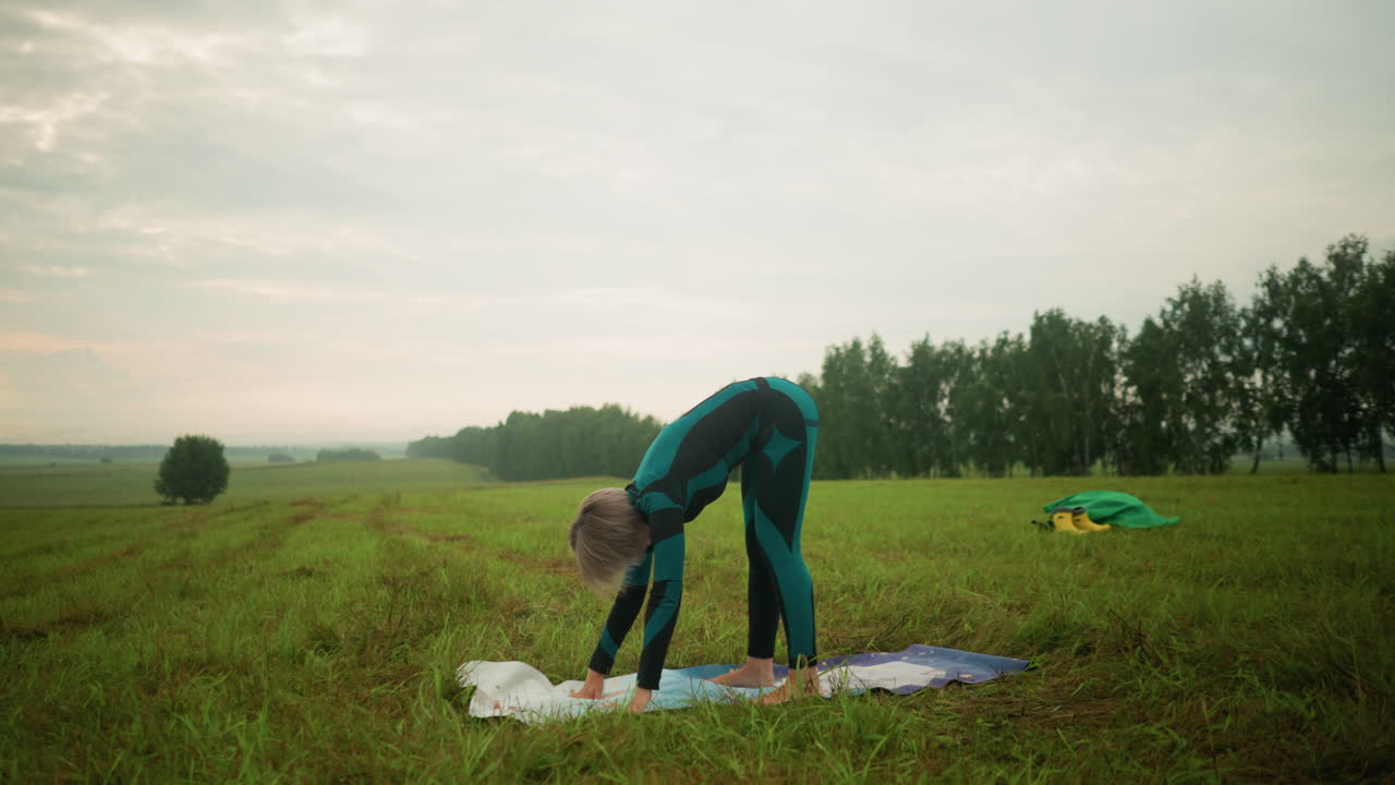 mujer en traje verde y negro realizando la postura del perro mirando hacia abajo, practicando yoga al aire libre en un campo cubierto de hierba bajo un cielo nublado, centrándose en el equilibrio, la flexibilidad y el estiramiento