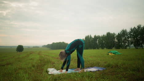 woman in green and black suit performing downward-facing dog pose, practicing yoga outdoors on a grassy field under a cloudy sky, focusing on balance, flexibility, and stretching