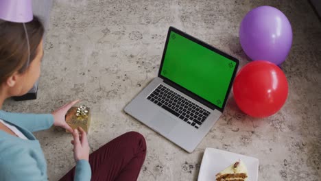 mujer sorprendida con sombrero de fiesta sosteniendo una caja de regalos mientras hace una videollamada en una computadora portátil en casa