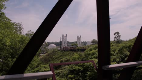 Un-Puente-De-Caballete-Metálico-Construido-Sobre-Una-Reserva-Natural-Con-Silos-Industriales-Al-Fondo