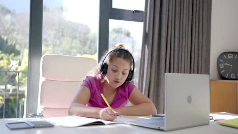 happy biracial girl using headphones and laptop for online school lesson at home