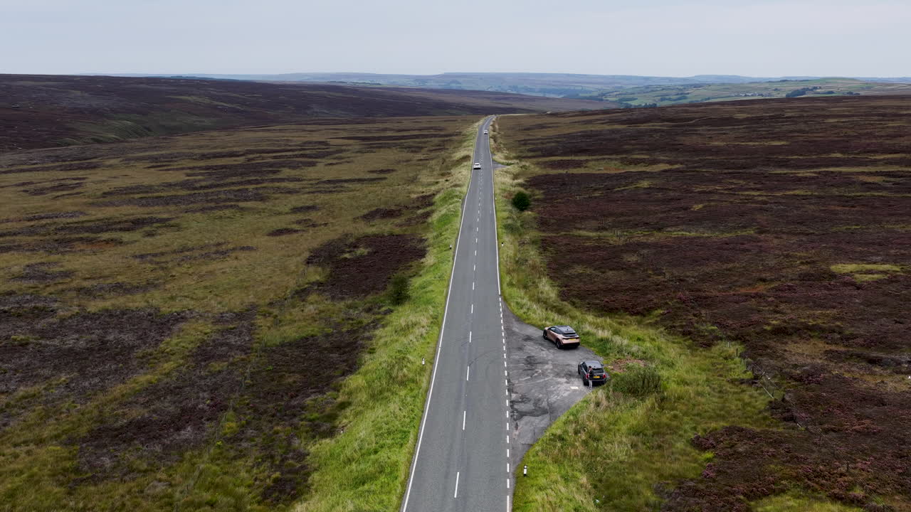 Wide aerial drone footage of a white Jaguar F‑Type convertible in the distance speeding along a rural West Yorkshire road, surrounded by fields and countryside scenery