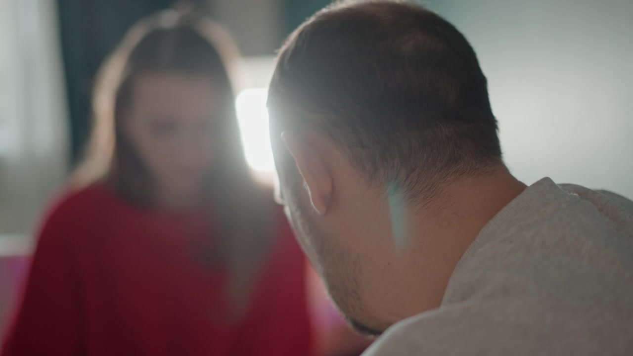 Escena doméstica cercana que muestra a un hombre adulto girando la cabeza hacia una mujer sentada con un suéter rojo, con un ligero movimiento de hombro, luz de contorno a contraluz, suave resplandor de ventana y un delicado destello de lente que sugiere una interacción atenta