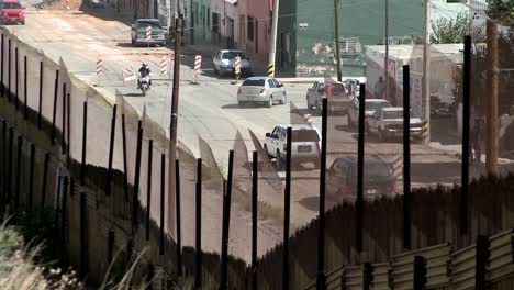 Cars-and-a-motorcycle-drive-down-a-street-a-pedestrian-crosses-and-other-people-stand-at-the-side