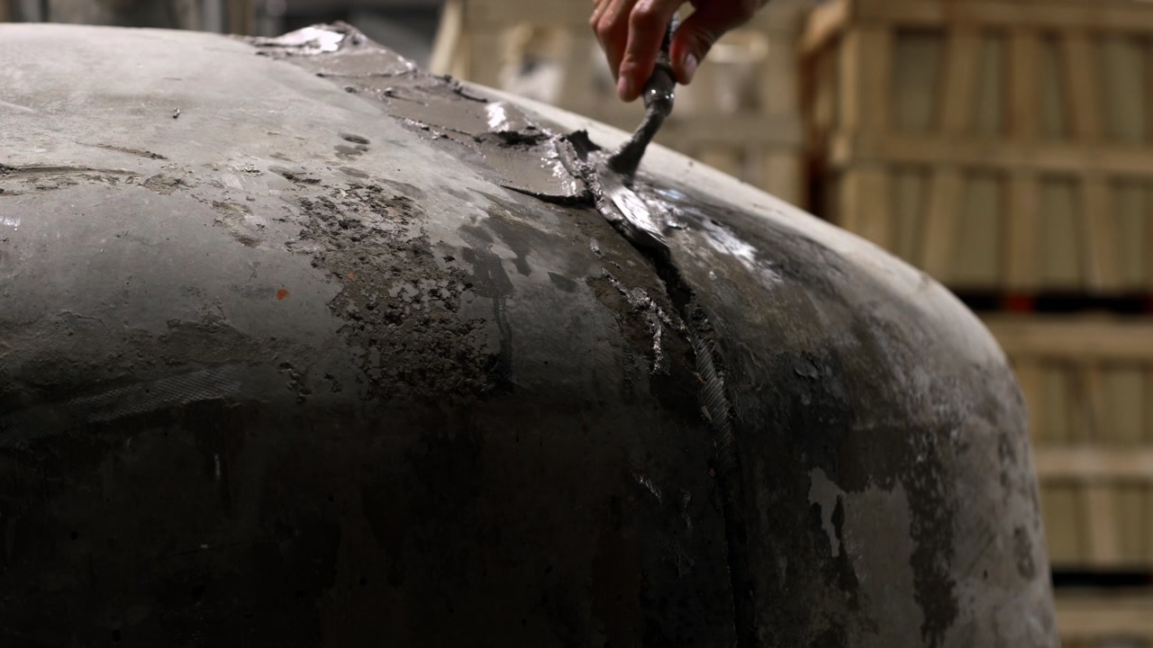 Static close-up shot of a worker’s hand applying wet grout along the seam of a curved pizza oven, highlighting texture and craftsmanship during construction.