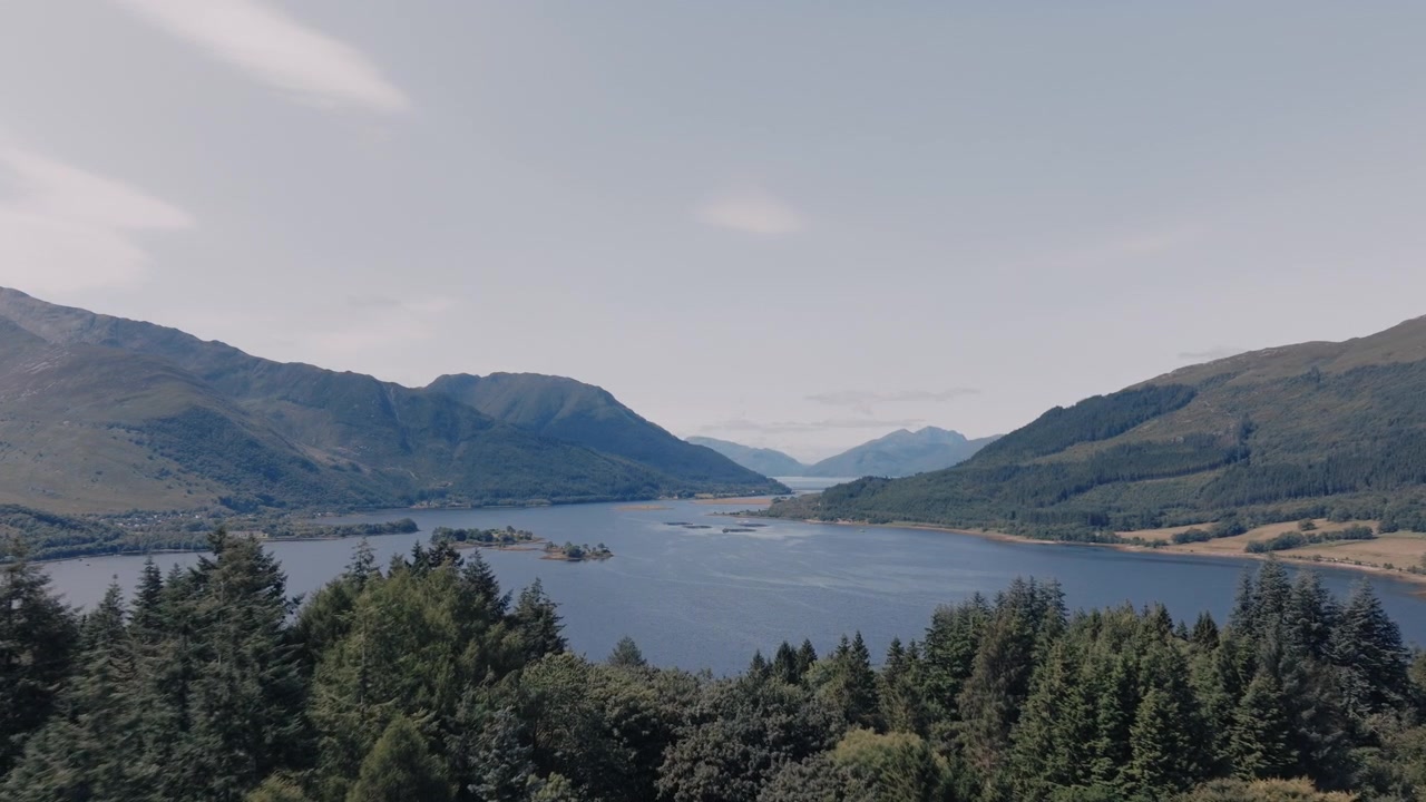 Luftfoto af en sø omgivet af fyrreskov med en lille holm og bjerge i baggrunden