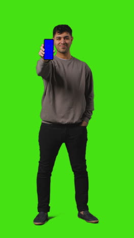 Vertical-Video-Full-Length-Shot-Of-Young-Man-Holding-Blue-Screen-Mobile-Phone-Standing-Against-Green-Screen