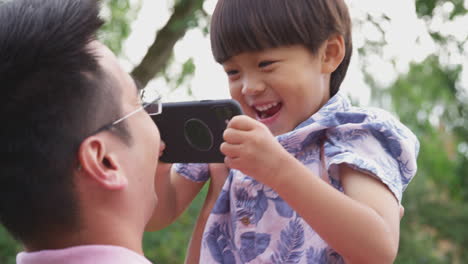 asian father playing with smiling son in garden as boy holds  mobile phone