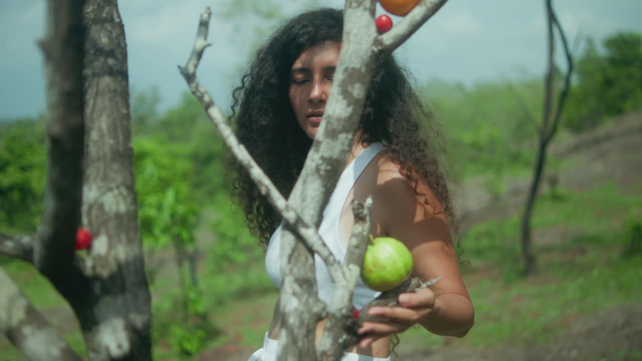 A woman in white stands near a tree with fruits, surrounded by green vegetation