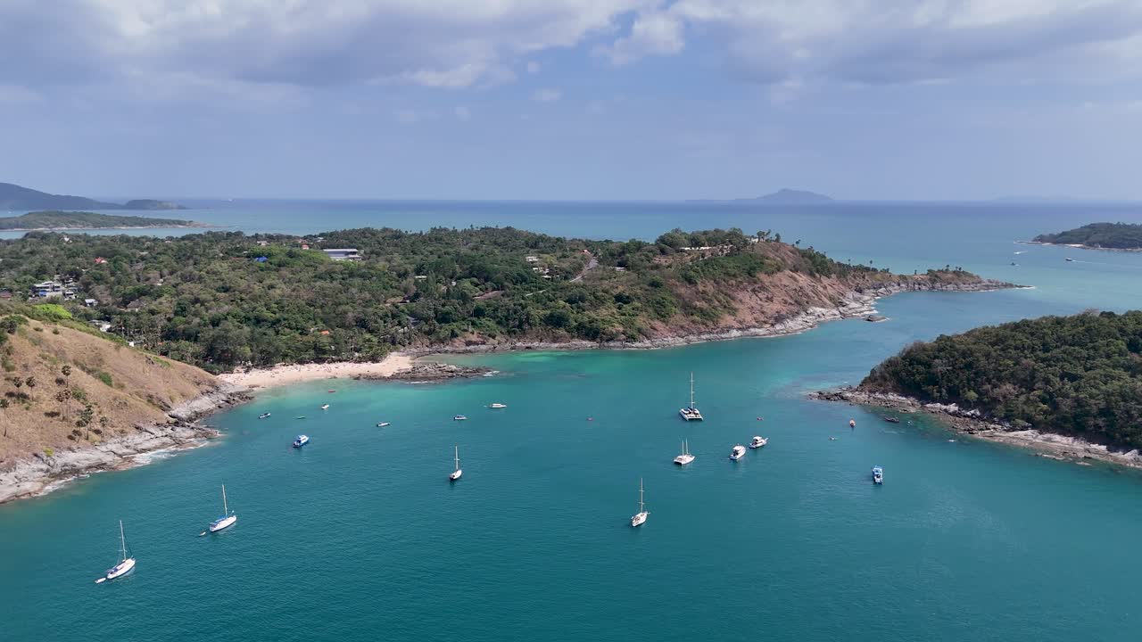 드론 영상은 푸켓의 멋진 해안선을 포착합니다. 푸르키즈색 물, 울창한 초록색, 그리고 은 하늘 아래 정박한 보트입니다.