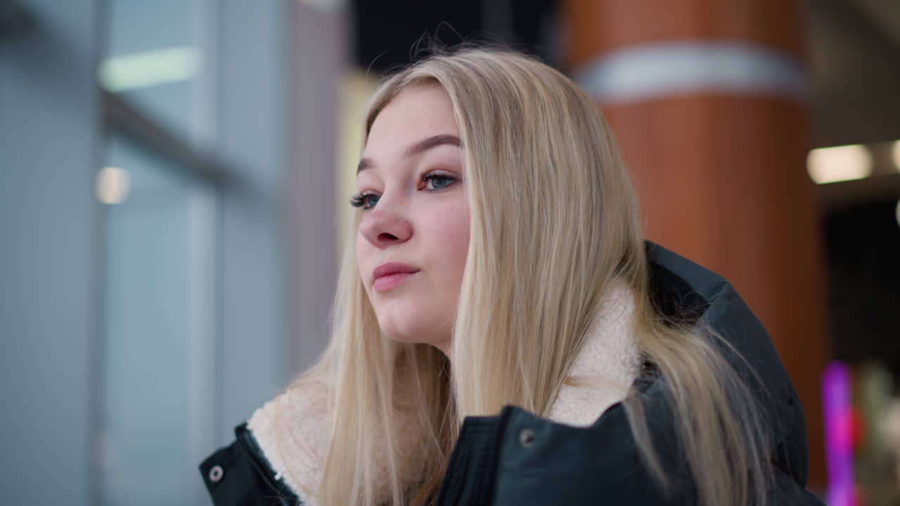 dama elegante con cabello rubio largo y sonrisa cálida mirando a su alrededor en el centro comercial, ella está usando una elegante chaqueta de invierno, de pie cerca de la barandilla de vidrio con fondo borroso