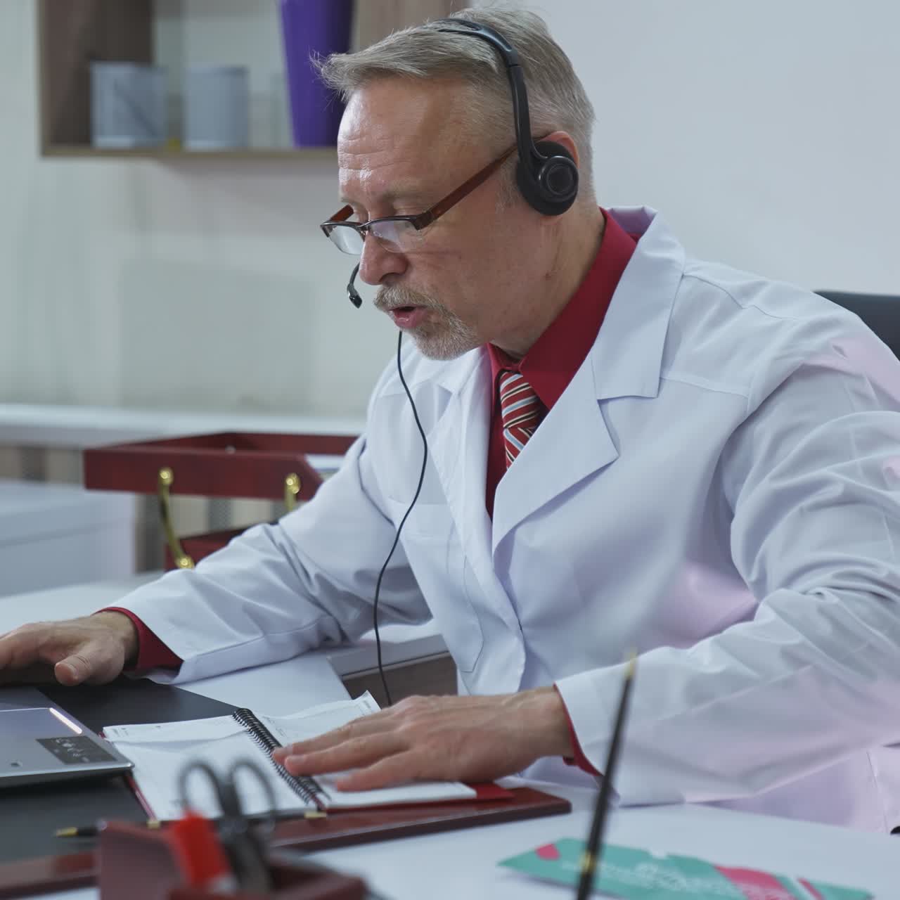 Old doctor in medical office working online. Professional male doctor in glasses and medical coat giving a consultation in front of laptop. Telemedicine. Coronavirus.