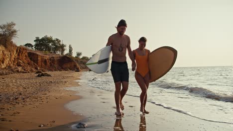 un hombre rubio en pantalones cortos negros camina y conversa con su novia en un traje de baño naranja, se toman de la mano y llevan tablas de surf mientras caminan a lo largo de la playa de arena rocosa por la mañana al amanecer