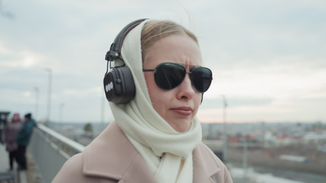 都会の女性が屋外で音楽を楽しんでいる。サングラスとコートを身につけ、冬の景色を眺めながらくつろぐ。バルコニーから冬の街並みを眺めつつ、音楽をじっくりと味わっている。