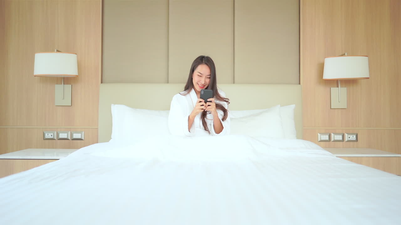 목욕 가운을 입은 한 젊은 여성이 고급스러운 호텔 침대에 앉아 스마트폰으로 전 세계에 문자 메시지를 보내고 있습니다.