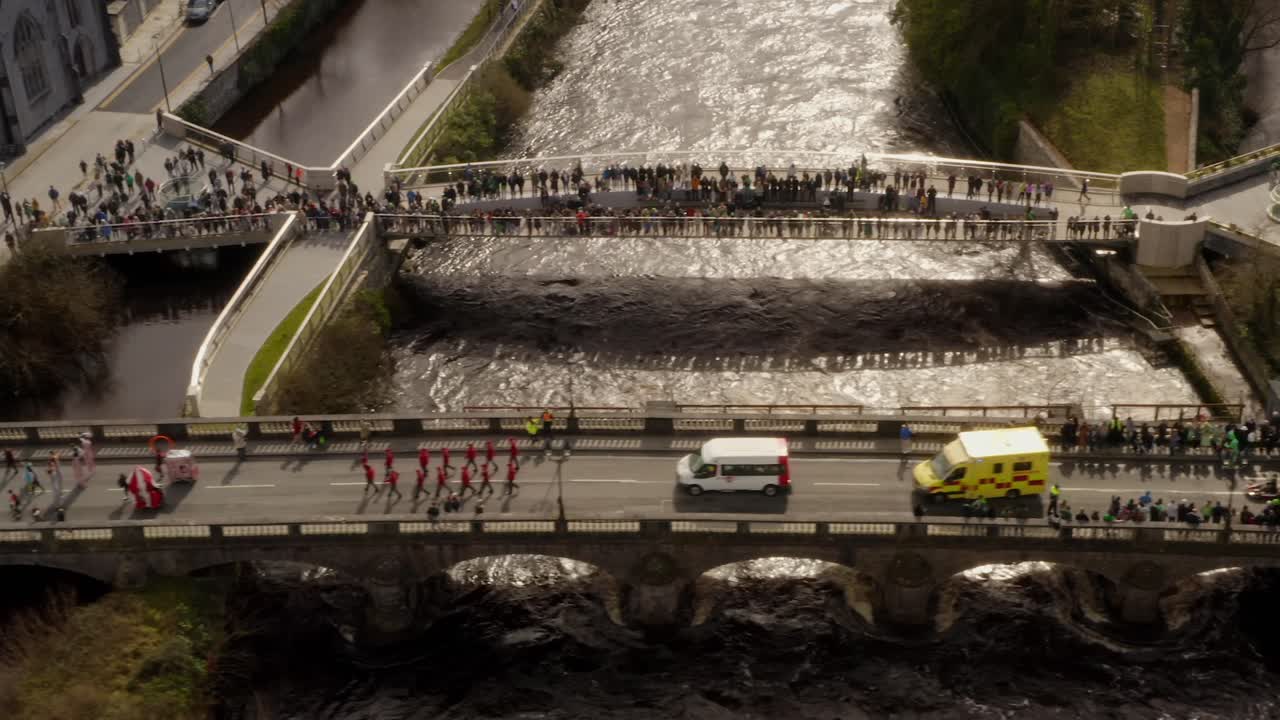 Aerial tilt down follows parade goers crossing bridge as families watch from Salmon Weir in Galway