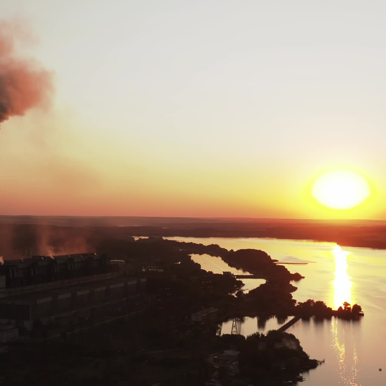 Silhouette of a factory near the river at sunset. Dark emissions from chimneys exhaust into the atmosphere against the orange setting sun. Ecology in danger