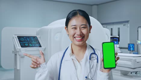 Close Up Of Asian Woman Doctor Holding Green Screen Smartphone With Stethoscope And Pointing To Side While Standing With Mri Machine In The Hospital