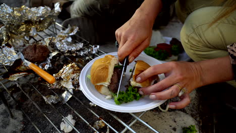 Weibliche-Hände-Schneiden-Burger-Draußen-Am-Kamin-In-Zwei-Hälften